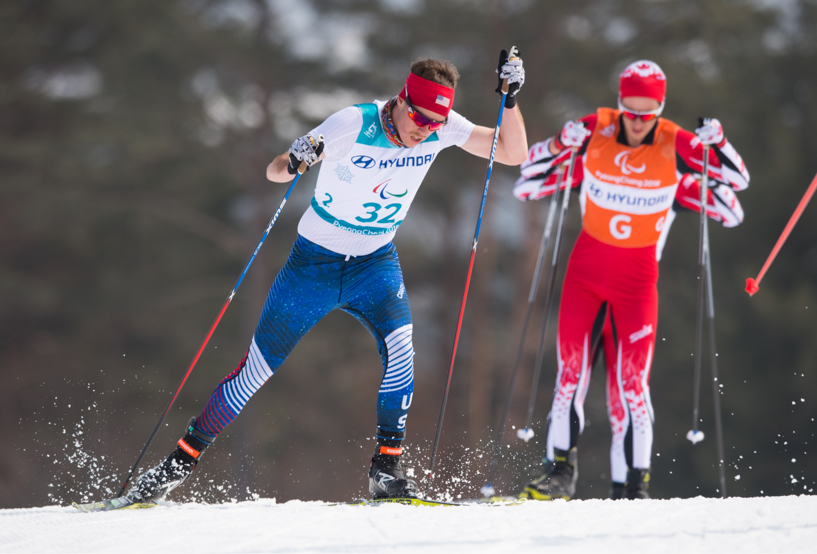 A front view of Jacob skiing in the Men's 2018 Paralympic 20 kilometer, Visually Impaired, Cross Country Skiing. Jacob is wearing a Team USA spandex race suit, red headband and sunglasses. He's also wearing a race bib with a number “32” on the front. In the background, slightly blurred, are a competing skier and guide racing for Team Canada in red race suits.