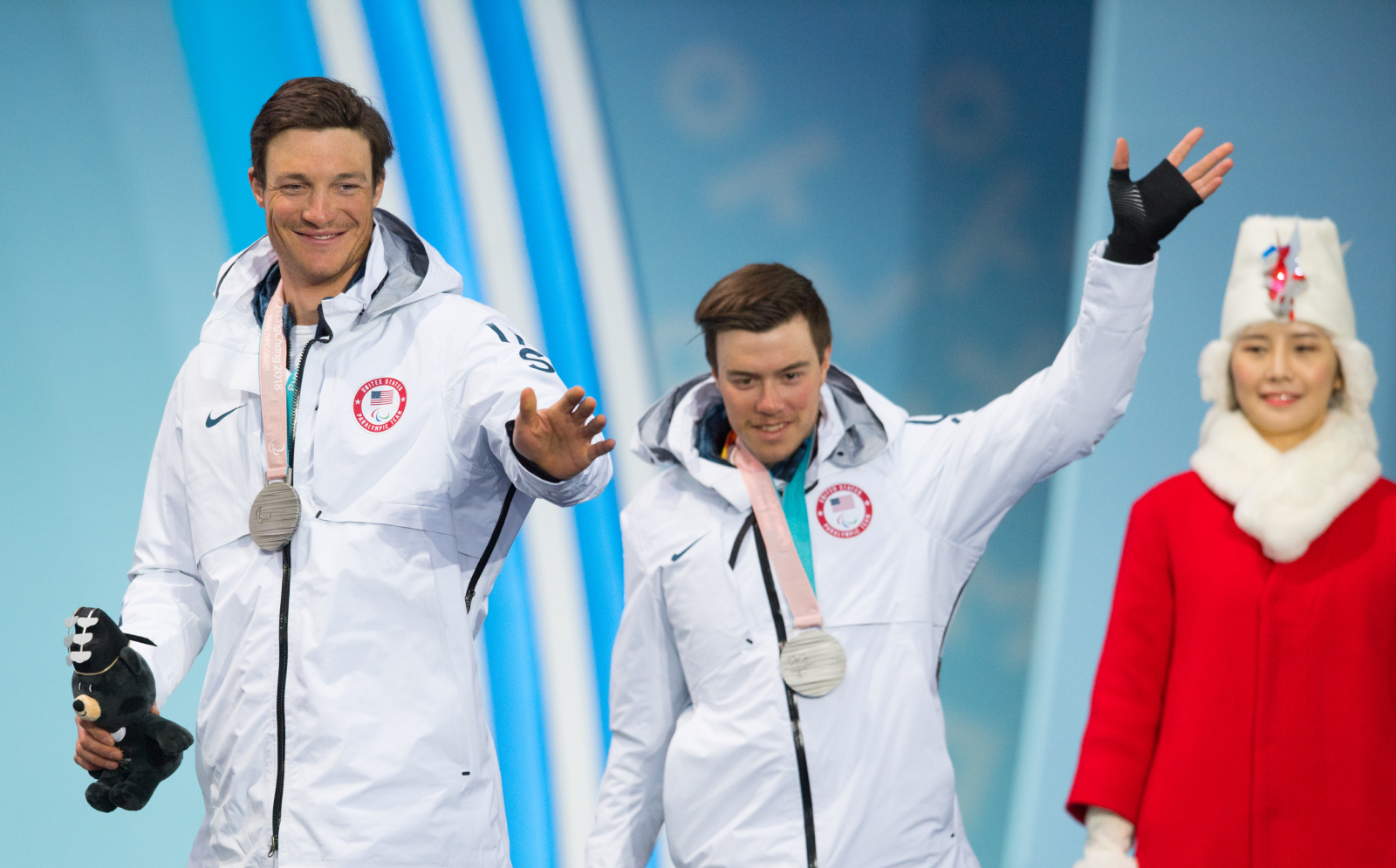 Jacob (right) and guide Sawyer Kesselheim (left), at the medal ceremony for the Men's 2018 Paralympic 10 kilometer, Visually Impaired, Cross Country Skiing. They are standing side by side, smiling, waving, with silver medals around their necks. They're wearing long, white Team USA jackets, and both holding a stuffed bear, the Pyeongchang Paralympic mascot named “Bandabi.” In the background stands one of the ceremonies medal hosts, wearing a red overcoat, white fur scarf and white hat. 